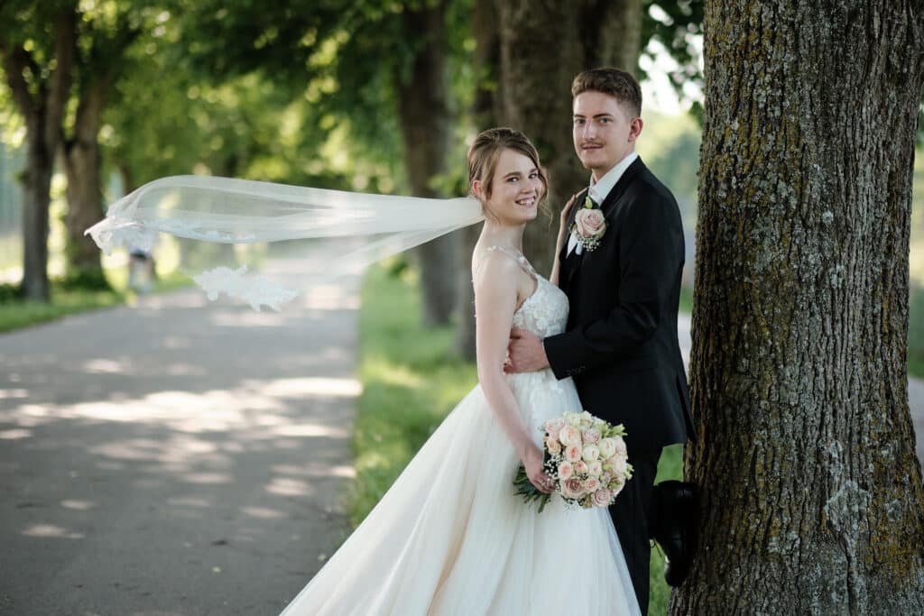 Brautpaar am Hochzeitstag im Sonnenschein unter einer Allee – Braut mit fliegendem Schleier und Rosenstrauß, Bräutigam im schwarzen Anzug